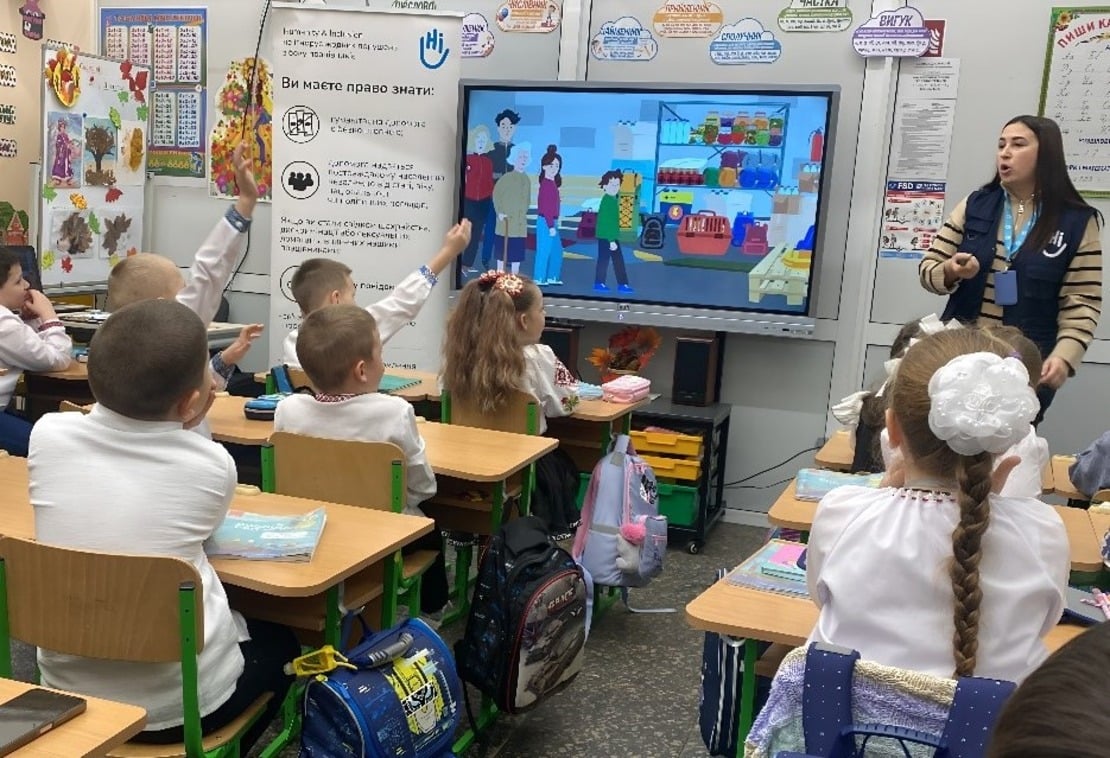 Kinderen zitten aan hun bureau in een klaslokaal voor een scherm waarop educatieve beelden te zien zijn van een Oekraïens gezin in hun dagelijks leven om veilig gedrag bij gevaar uit te leggen. Op het schoolbord staat een medewerker van Handicap International die de beelden aan hen beschrijft. 