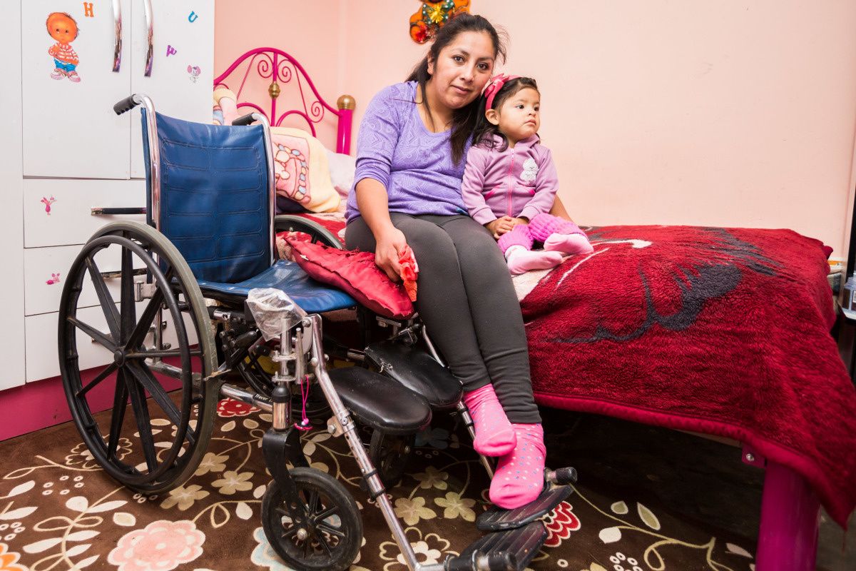 Een vrouw en haar dochtertje zitten op bed, met daarnaast een rolstoel