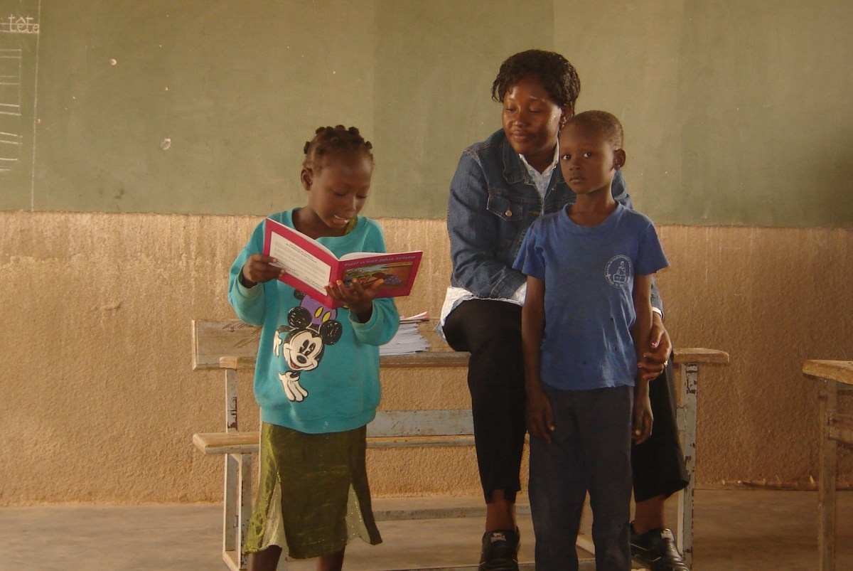 Jonge vrouw uit Burkina Faso zit vooraan op een bank in de klas terwijl ze een jongen dichtbij haar houdt en luistert hoe een ander kind voorleest uit een boek vooraan in de klas.