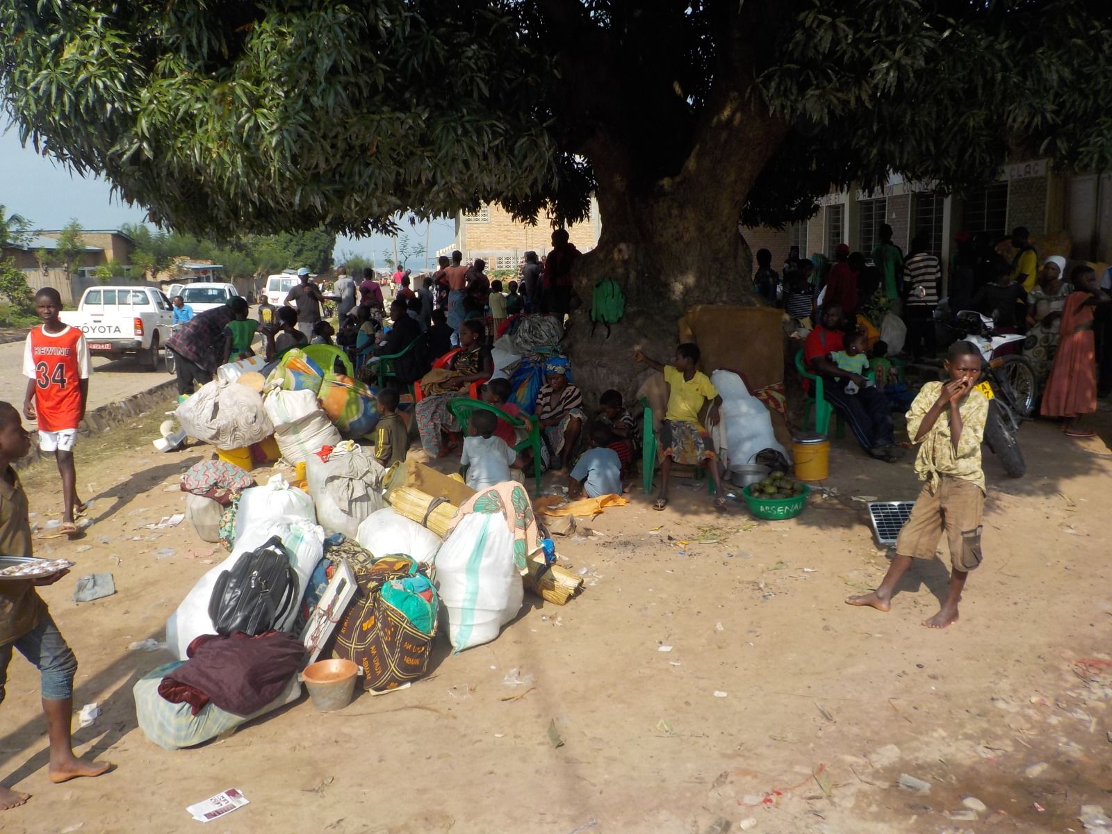 Een groep Congolese vluchtelingen zit in de schaduw van een grote boom, met de zakken die ze konden dragen naast hen.