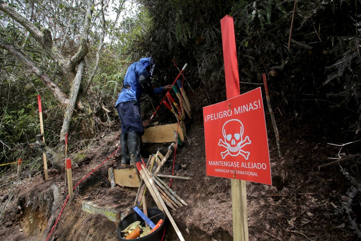 Un démineur au travail sur un terrain escarpé dans une forêt. A l'avant-plan, un panneau rouge avec le texte 