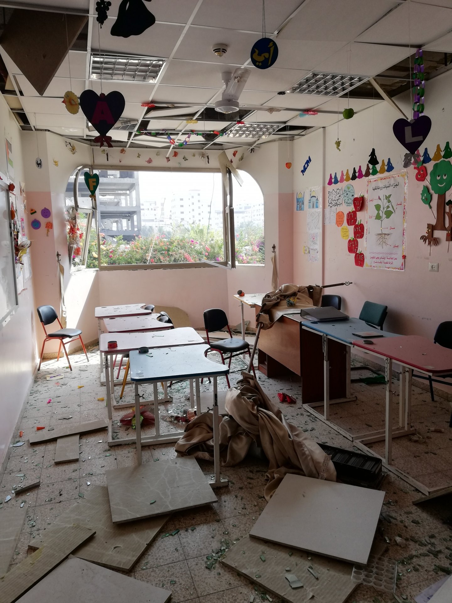 Het interieur van een beschadigde klas: gebroken vensters, glas op de grond, losgekomen pleister aan het plafond.