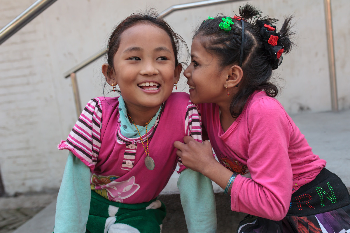 Twee Nepalese meisjes zitten neer, waarbij de ene iets in het oor fluistert van het andere