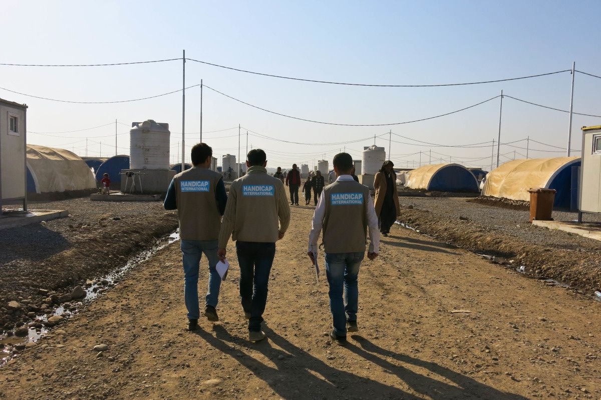 Drie mannen in vest van Handicap International wandelen tussen de tenten van het kamp van Khazer