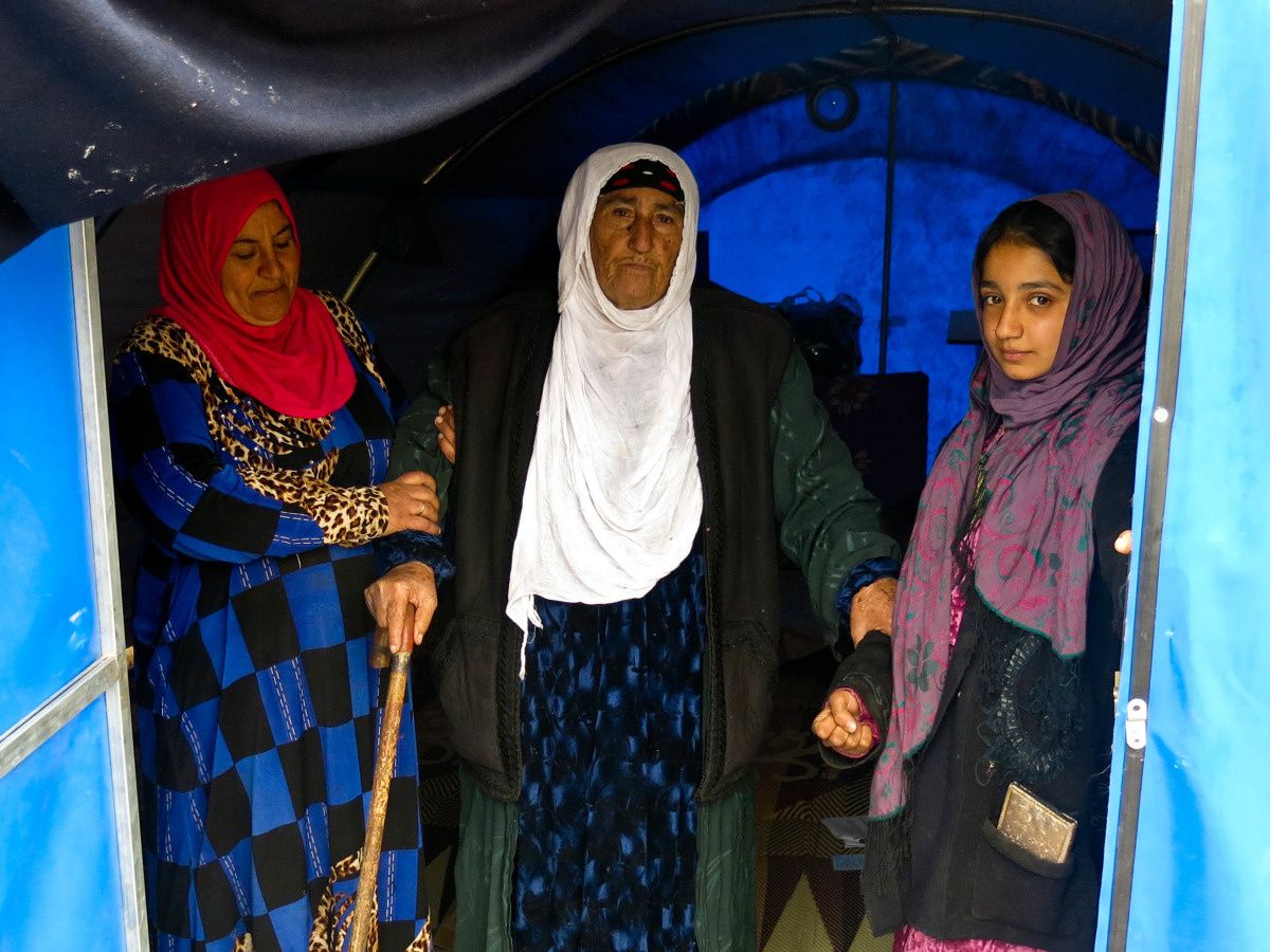 Drie gesluierde vrouwen aan de ingang van een tent, waarbij de oudste op krukken steunt