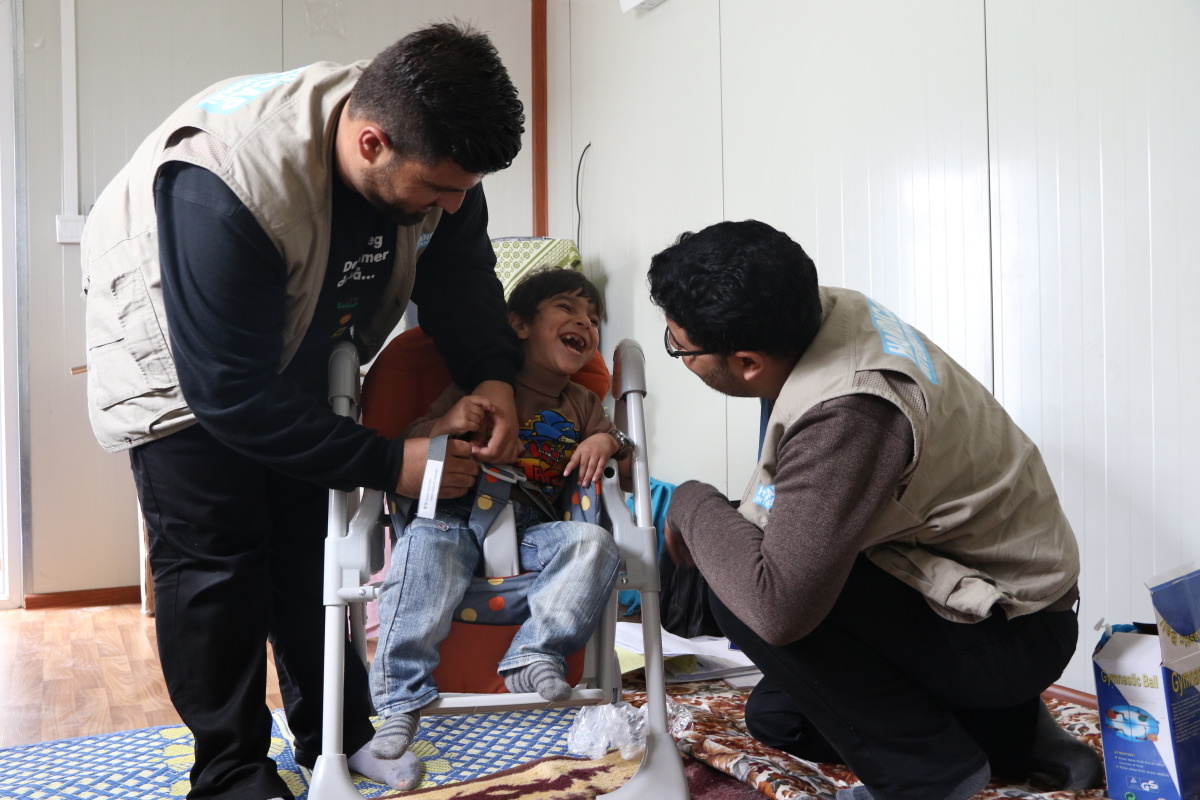 Twee mannen in vest van Handicap International buigen zich rond Sofian, die schaterlacht terwijl hij in zijn rolstoel zit.