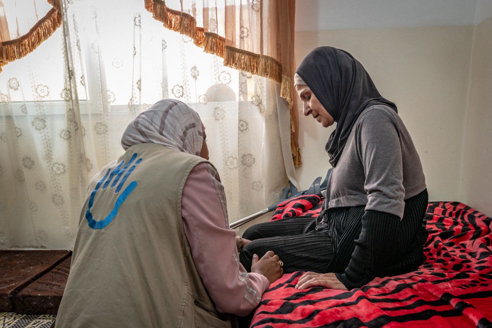 Une femme assise sur son lit. Une kiné de Handicap International examine sa jambe.