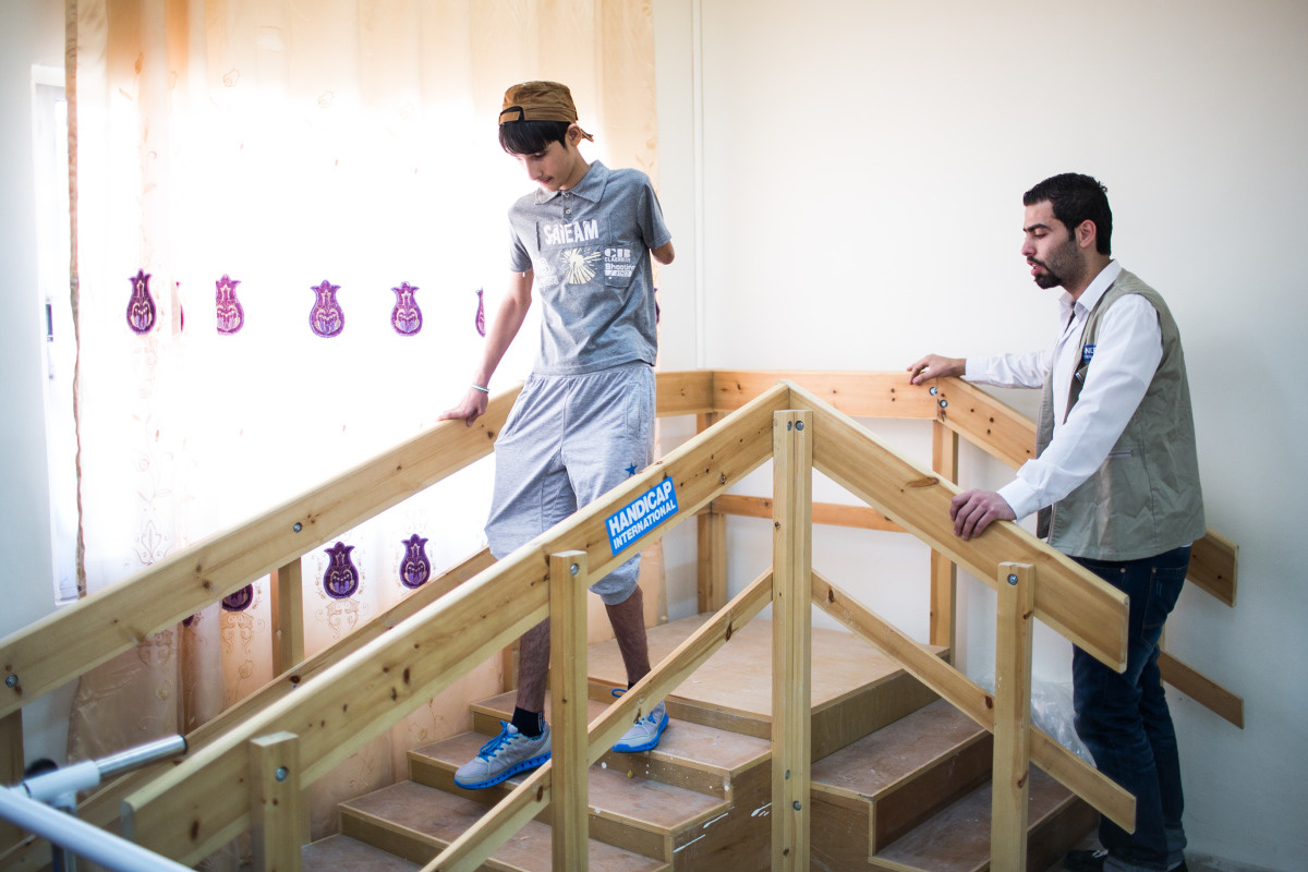 Een jongen zonder linkerarm en met een been met externe fixator daalt een houten loopbrug af in een revalidatiezaal onder begeleiding van een man in een jasje van Handicap International