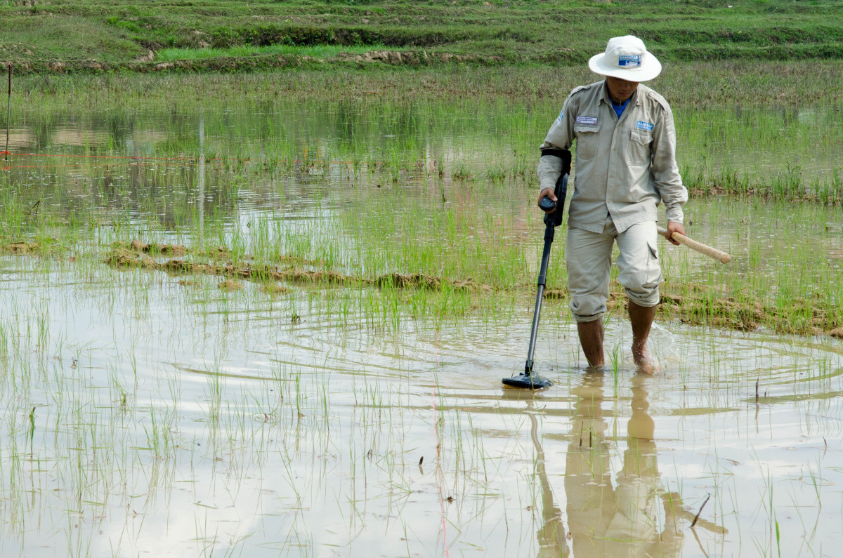 Een man in ontmijningsoutfit loopt door een veld met enkelhoog water en doorzoekt het met een metaaldetector