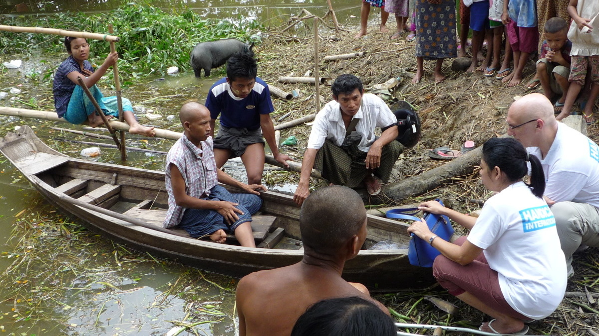 hulpverleners in t-shirt van handicap international knielen neer en discussiëren bij een houten bootje dat aanmeert met een myanmarees als passagier