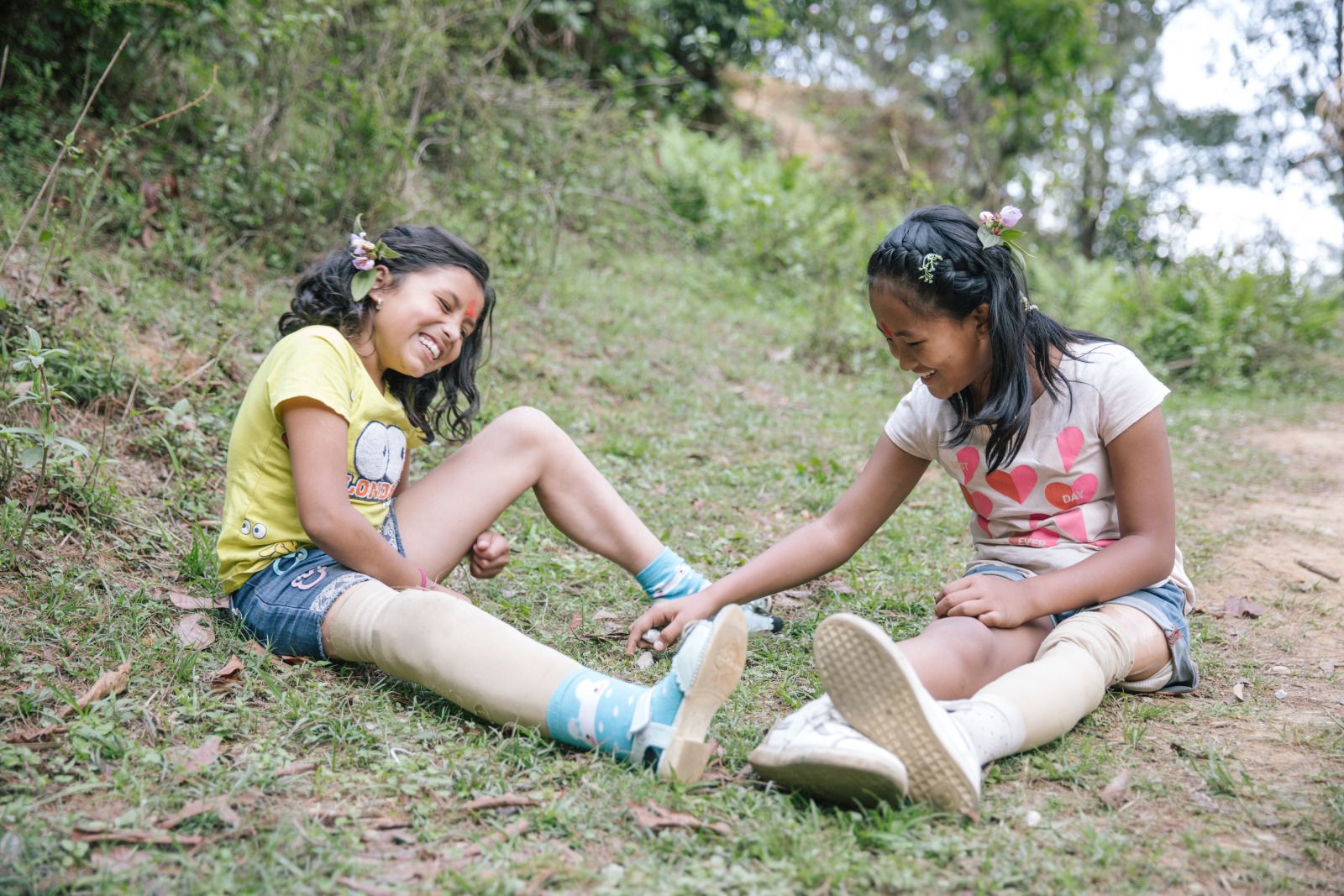 Twee jonge Nepalese meisjes van ongeveer 12 jaar, allebei met een prothese, praten zittend in het gras.