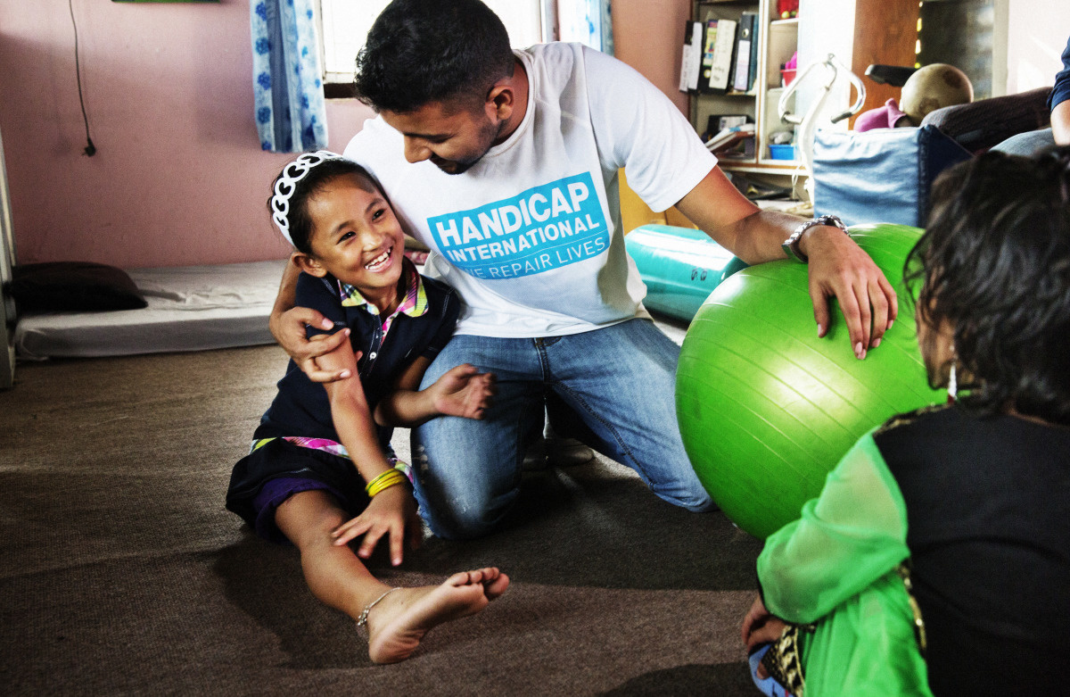 Een kinesitherapeut in T-shirt van Handicap International heeft een grote bal voor kine-oefeningen vast en omarmt de lachende Khembro, een meisje met slechts één been. 