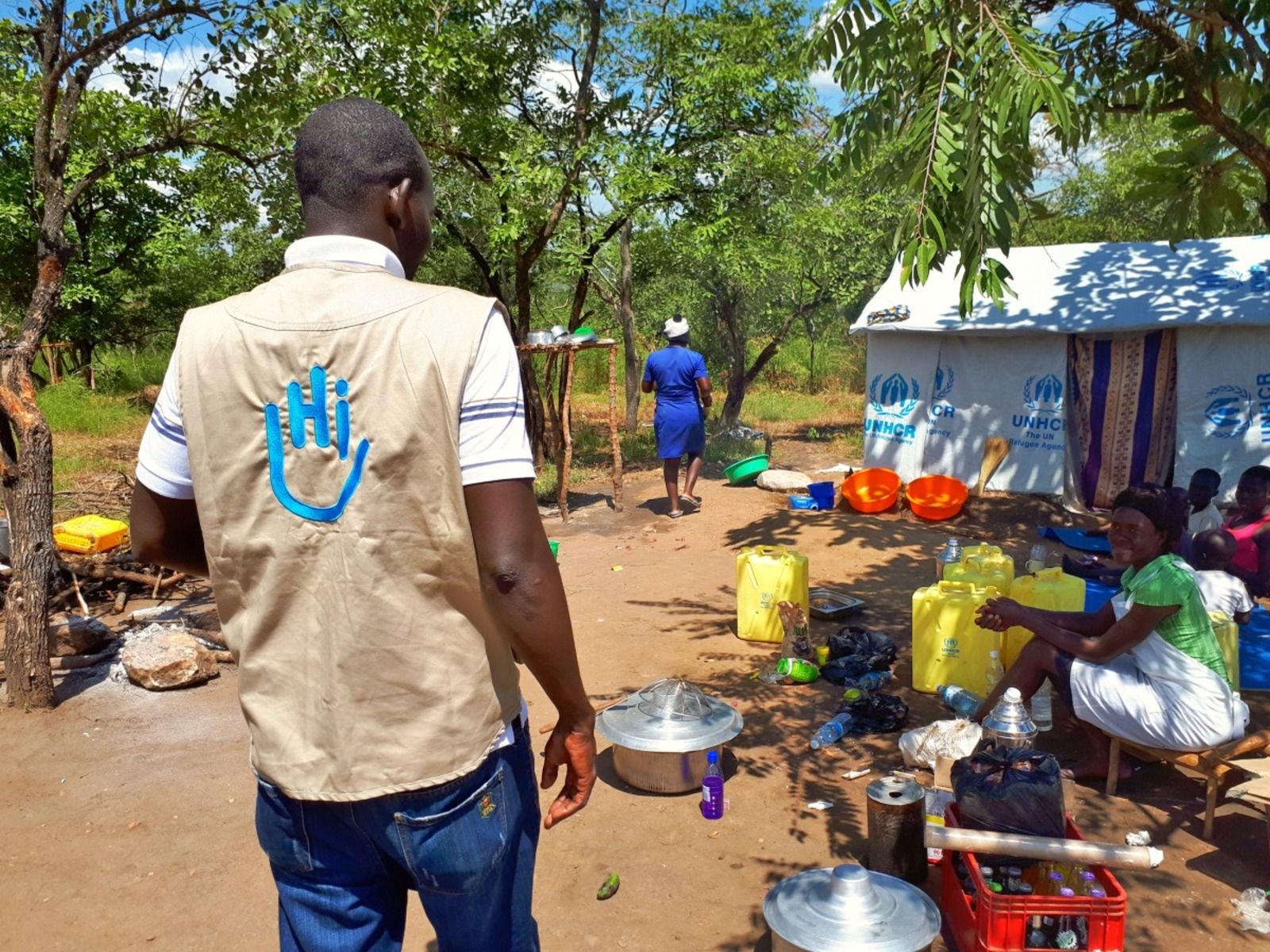 Foto van een Afrikaanse man. Op de achtergrond zit een vrouw naast grote jerrycans met water en een tent gemaakt uit zeilen