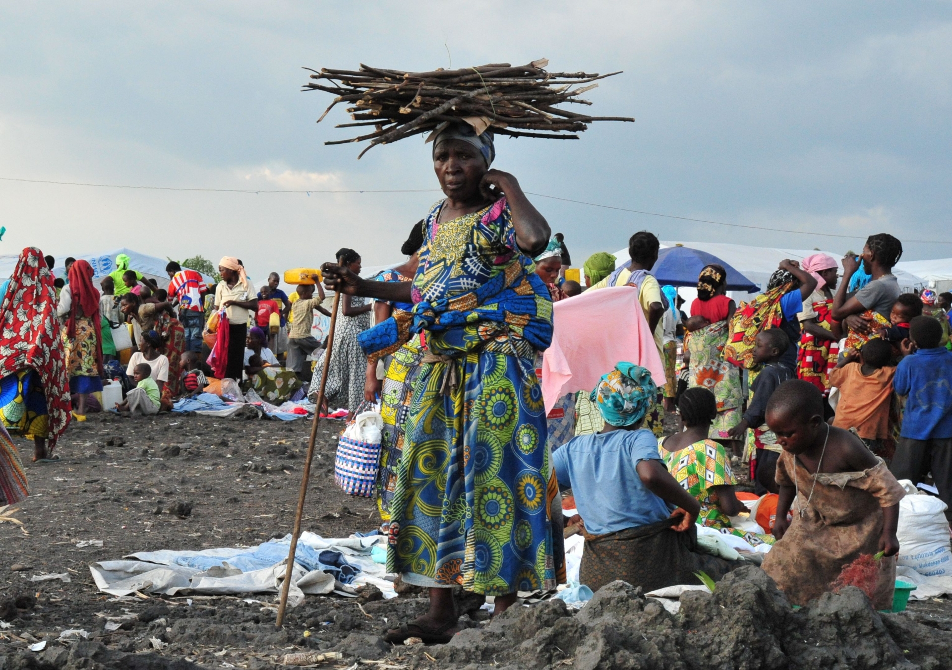 Vluchtelingenkamp in Congo. Vrouw van middelbare leeftijd draagt een stapel takken op haar hoofd. Tientallen mensen rondom zitten neer of staan recht. 