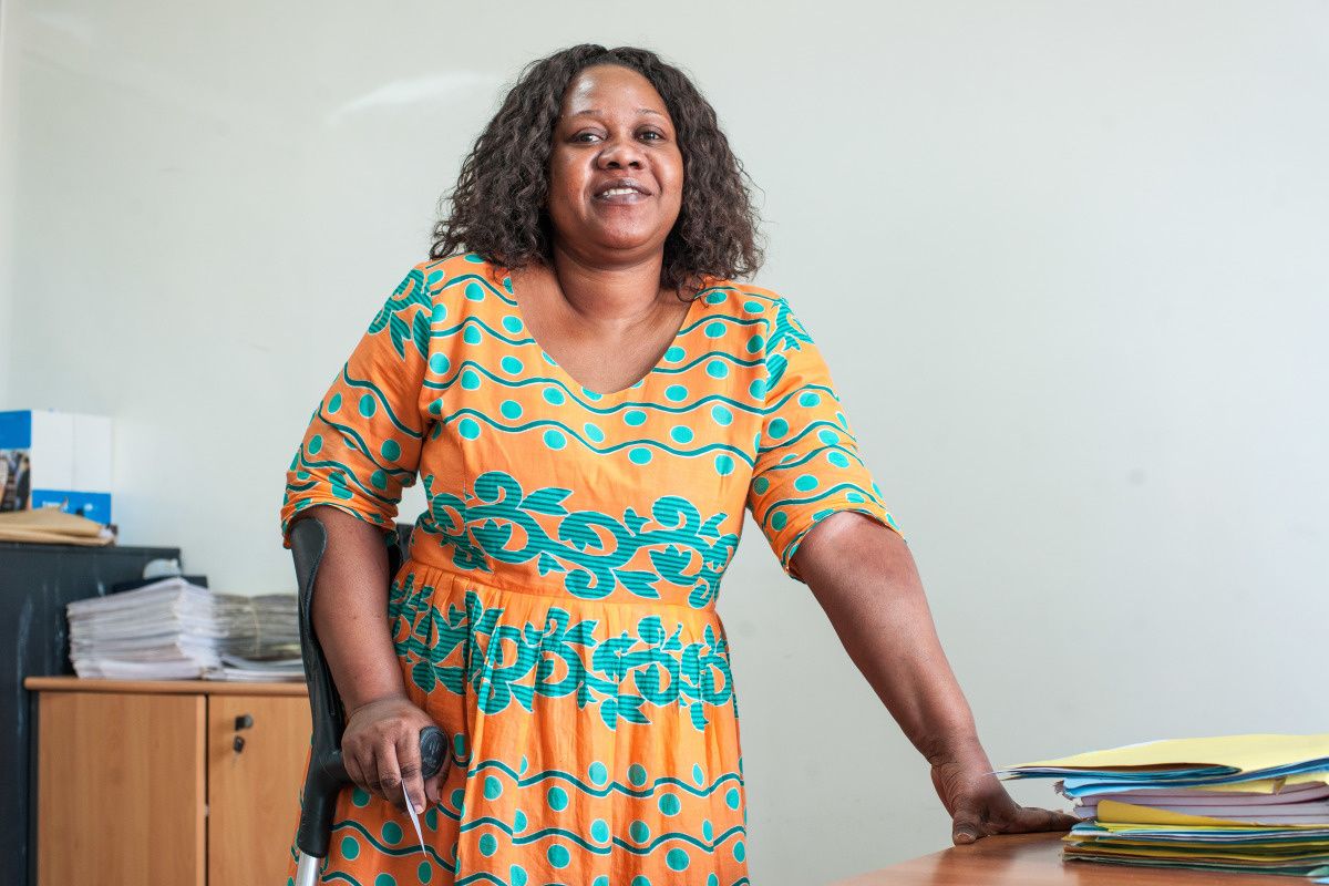 Une femme dans une robe jaune avec des motifs bleus, debout à côté d'un bureau. Sa main gauche est posée sur le bureau, son bras droit s'appuie sur une béquille