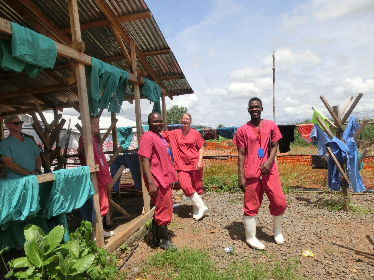 Vier medewerkers van Handicap International in Sierra Leone glimlachen voor de foto, terwijl ze bij een gebouw staan waar groene verzorgingsoutifts hangen te drogen