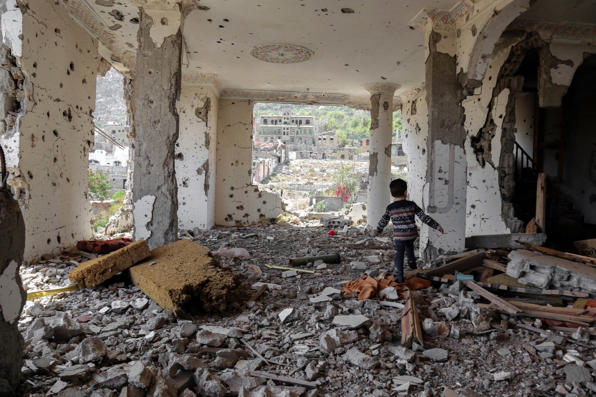 Un enfant de +/- 6 ans, de dos, marche au milieu des décombres d'un bâtiment. Le batiment est encore debout mais le sol est couvert de débris, il n'y a plus une seule vitre.