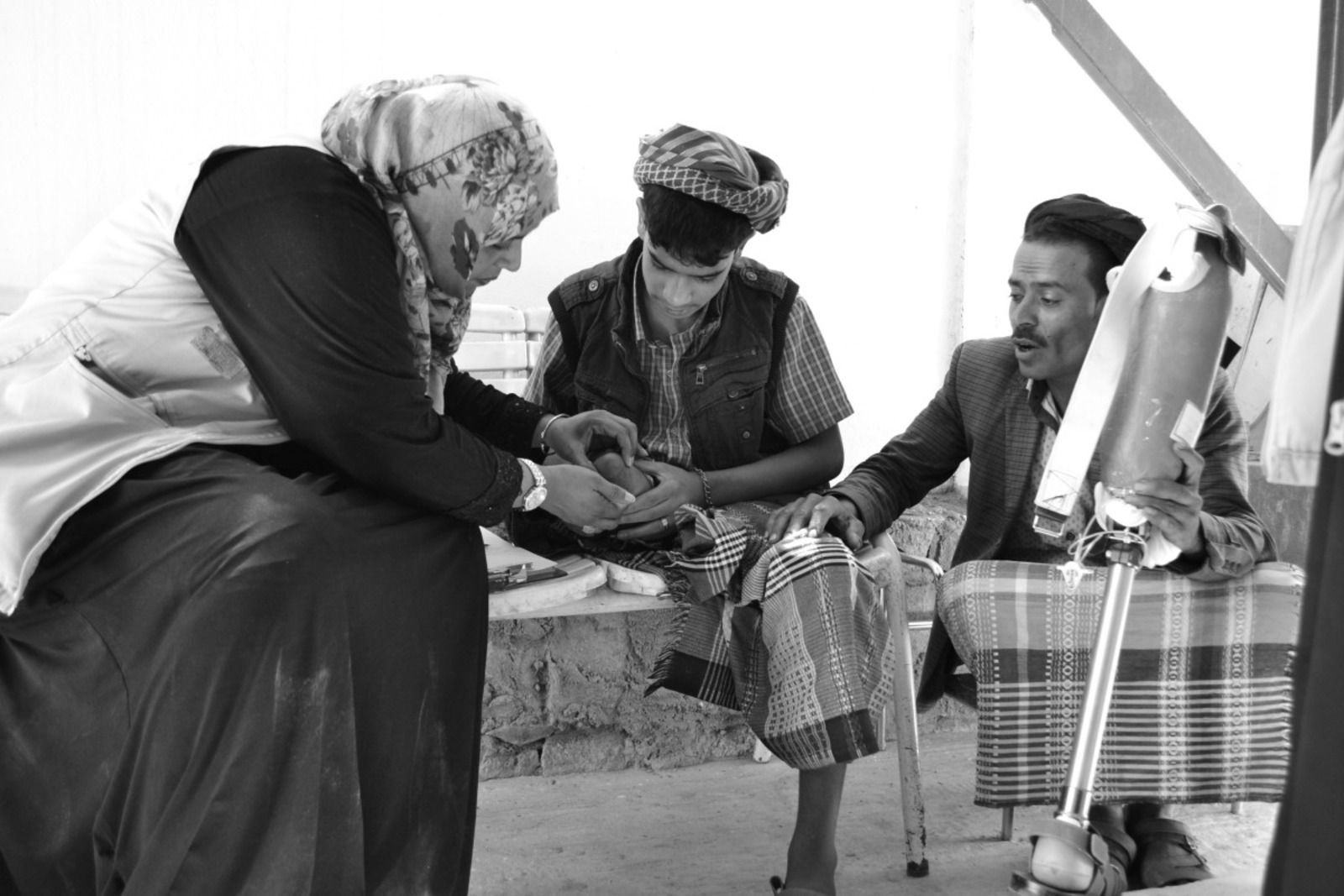 Photo en noir et blanc, une dame voilée employée de HI se penche vers un adolescent amputé  de la jambe droite. De l'autre côté, un homme tient une prothèse.