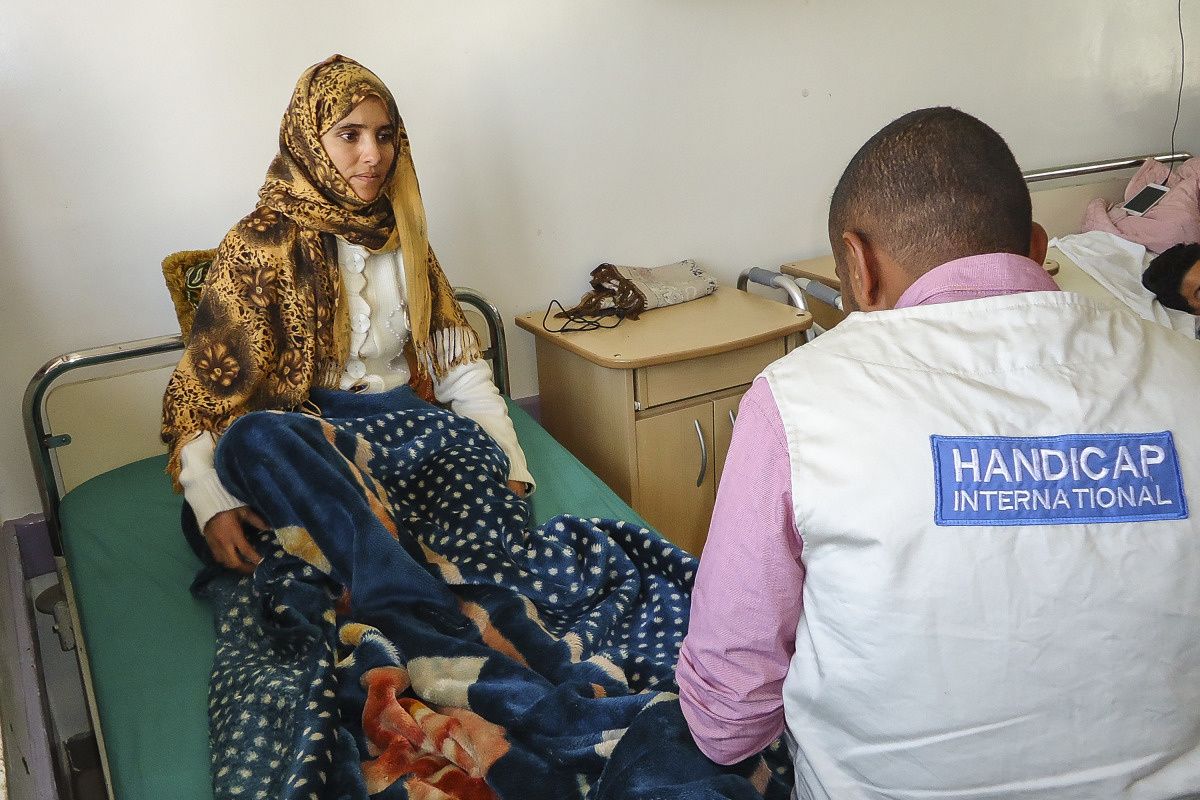 Bushra, une jeune femme, assise sur un lit pendant qu'un kinésithérapeute de HI, assis de dos, l'examine.